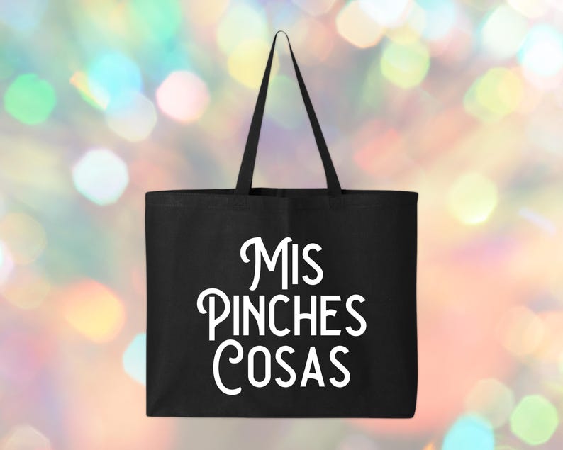 May include: A black tote bag with the words "MIS PINCHES COSAS" in white script. The bag has a rectangular shape with a handle extending from the top. The background is a colorful bokeh effect.