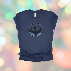 May include: Navy blue t-shirt featuring a circular design with a black bird silhouette inside a light blue circle. The bird is depicted with outstretched wings. The shirt is made of a soft material.