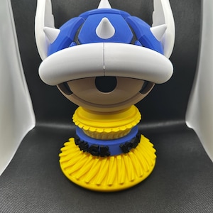 May include: A 3D-printed sculpture of a blue Koopa shell with white spikes and accents. The sculpture has a yellow base with the word "WACK" in black letters. The shell is a recognizable element from a popular video game.