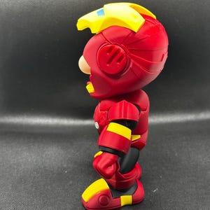 Puede incluir: Vista lateral de una figura de acci&oacute;n roja y amarilla. La figura tiene un casco rojo con detalles amarillos, un tocado amarillo y una armadura roja y amarilla. La figura est&aacute; sobre una superficie negra.
