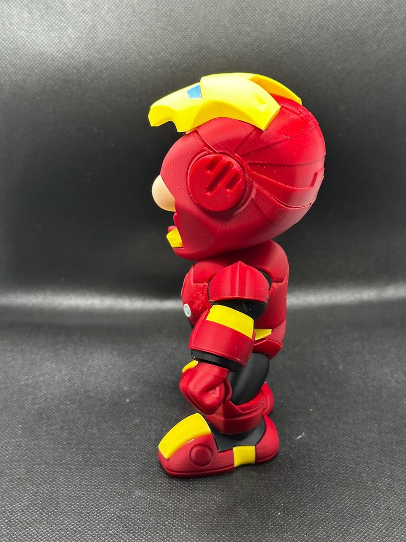 Puede incluir: Vista lateral de una figura de acci&oacute;n roja y amarilla. La figura tiene un casco rojo con detalles amarillos, un tocado amarillo y una armadura roja y amarilla. La figura est&aacute; sobre una superficie negra.