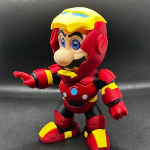Puede incluir: Una figura de acci&oacute;n roja, amarilla y negra de Mario con un traje de Iron Man. La figura tiene un casco amarillo con detalles azules, una placa pectoral roja con un c&iacute;rculo azul y botas rojas con detalles amarillos. La figura est&aacute; posando con un brazo extendido.