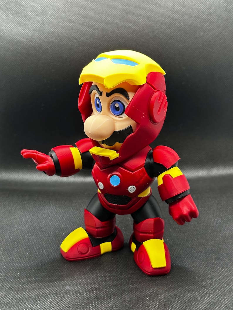 Puede incluir: Una figura de acci&oacute;n roja, amarilla y negra de Mario con un traje de Iron Man. La figura tiene un casco amarillo con detalles azules, una placa pectoral roja con un c&iacute;rculo azul y botas rojas con detalles amarillos. La figura est&aacute; posando con un brazo extendido.