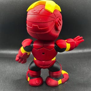 Puede incluir: Figura de acci&oacute;n roja y amarilla con una cabeza en forma de casco, brazos extendidos y un dise&ntilde;o detallado. La figura tiene detalles en negro y est&aacute; sobre un fondo oscuro.