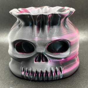 Puede incluir: Un recipiente con forma de calavera impreso en 3D con una parte superior en forma de bolsa. El recipiente es principalmente negro con vetas rosas y moradas. La calavera presenta cuencas de los ojos, una cavidad nasal y una hilera de dientes.