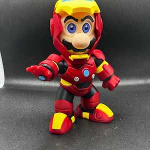 Puede incluir: Una figura de Mario con un traje de Iron Man. La figura es roja, amarilla, negra y azul. El casco es amarillo con detalles rojos. La figura tiene ojos azules y bigote. La figura est&aacute; sobre una superficie oscura.