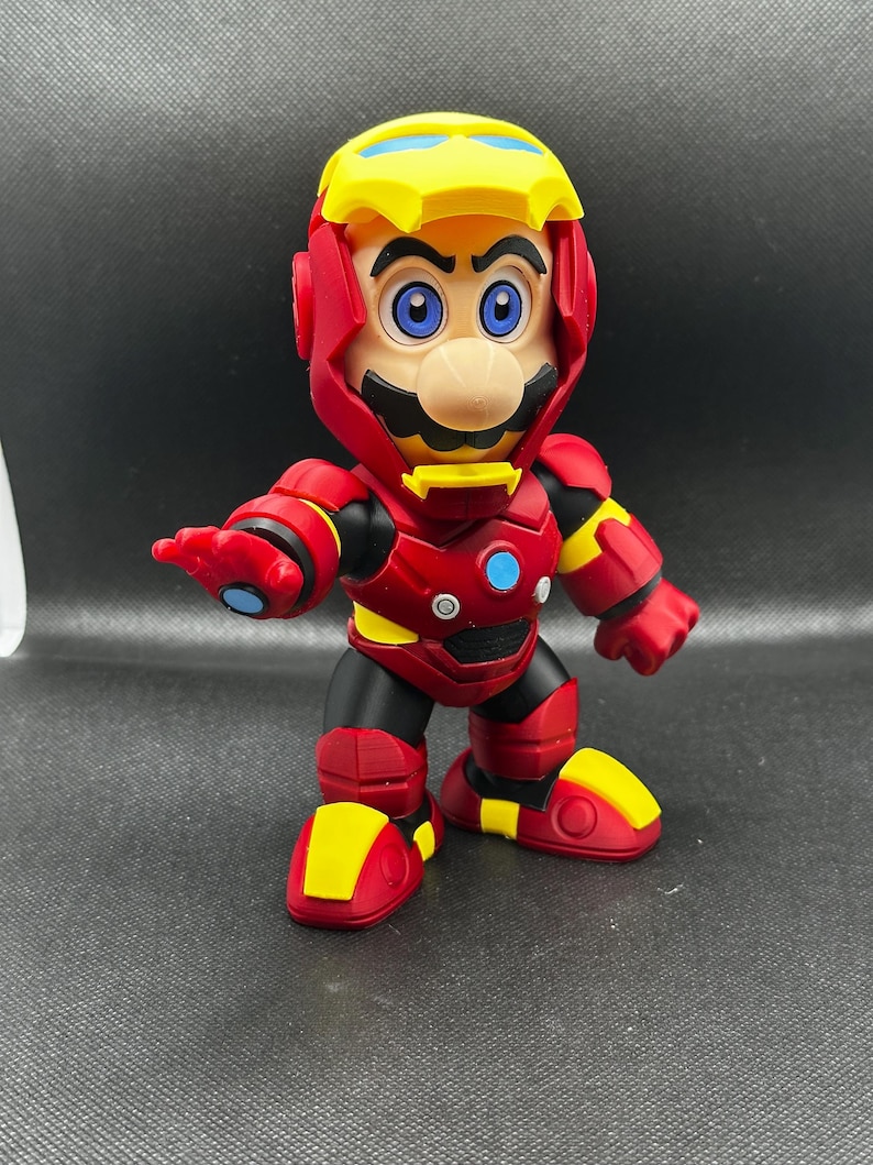 Puede incluir: Una figura de Mario con un traje de Iron Man. La figura es roja, amarilla, negra y azul. El casco es amarillo con detalles rojos. La figura tiene ojos azules y bigote. La figura est&aacute; sobre una superficie oscura.