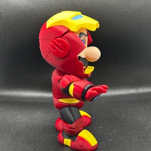 Puede incluir: Una figura de acci&oacute;n roja y amarilla de Mario con un traje inspirado en Iron Man. La figura tiene un casco amarillo con detalles azules, una armadura roja y detalles amarillos en los pies y los brazos. La figura est&aacute; posando con un brazo extendido.