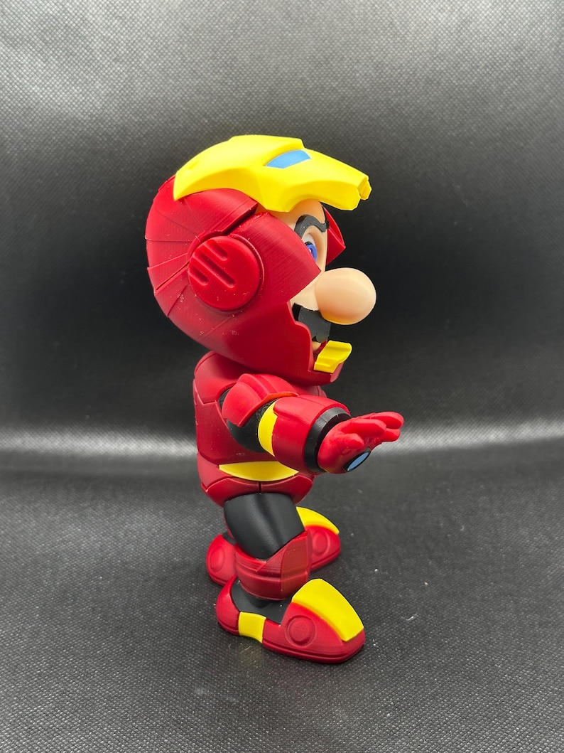 Puede incluir: Una figura de acci&oacute;n roja y amarilla de Mario con un traje inspirado en Iron Man. La figura tiene un casco amarillo con detalles azules, una armadura roja y detalles amarillos en los pies y los brazos. La figura est&aacute; posando con un brazo extendido.