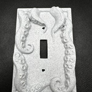Könnte beinhalten: Hellgraue, strukturierte Lichtschalterabdeckung mit detaillierten Reliefs von Oktopus-Tentakeln. Die Tentakel winden sich um die Kanten und umrahmen die Schalteröffnung und die Schraubenlöcher. Die Abdeckung hat ein gesprenkeltes Muster.