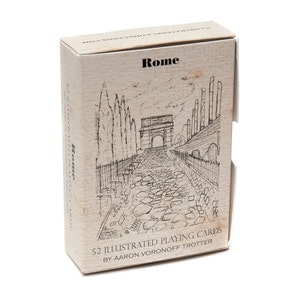 May include: A deck of playing cards with an illustration of a cobblestone street in Rome, Italy. The box is beige with black text that reads "Rome" and "52 Illustrated Playing Cards by Aaron Voronoff Trotter".