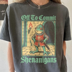 May include: A charcoal grey short-sleeve t-shirt featuring a graphic of a frog dressed as a medieval knight with a sword and torch. The text above the graphic reads Off To Commit and the text below reads Shenanigans in a light green font.