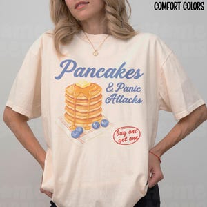 May include: An ivory-coloured t-shirt with the text "Pancakes & Panic Attacks" in blue script. The graphic features a stack of pancakes with butter, syrup, and blueberries. A red "buy one get one" circle is also visible.