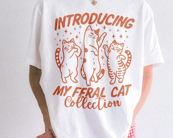 Könnte beinhalten: Ein weißes kurzärmeliges T-Shirt mit einem Grafikdruck in orangefarbener Tinte. Das Design zeigt drei Katzen und den Text "INTRODUCING MY FERAL CAT Collection", umgeben von kleinen Sternen. Das T-Shirt hat einen Rundhalsausschnitt und eine lockere Passform.