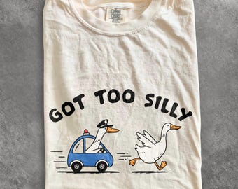 May include: An ivory short-sleeve t-shirt featuring a graphic of a white goose wearing a police hat driving a blue car, followed by another white goose running. The text "GOT TOO SILLY" is printed in black above the geese. The shirt has a crew neck.