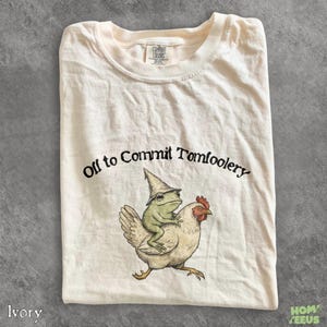 Könnte beinhalten: Ein elfenbeinfarbenes Kurzarm-T-Shirt mit einer Grafik eines grünen Frosches mit spitzem Hut, der auf einem weißen Huhn reitet. Der Text "Off to Commit Tomfoolery" ist in Schwarz über der Illustration gedruckt. Das Shirt hat einen Rundhalsausschnitt.