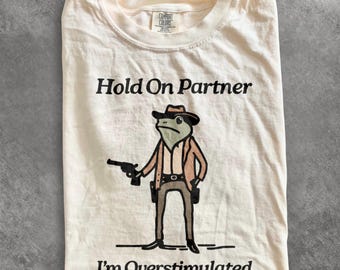 Puede incluir: Una camiseta de color marfil con un gráfico de una rana vestida de vaquero, sosteniendo una pistola negra. El texto sobre la rana dice Hold On Partner y el texto debajo dice I'm Overstimulated. La camiseta tiene cuello redondo.