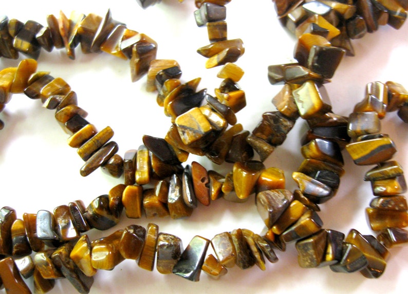 K&ouml;nnte beinhalten: Eine Str&auml;hne aus Tigerauge-Edelsteinkugeln, mit einer Mischung aus braunen, goldenen und schwarzen Farben. Die Kugeln sind unregelm&auml;&szlig;ig geformt und haben eine nat&uuml;rliche, raue Textur.