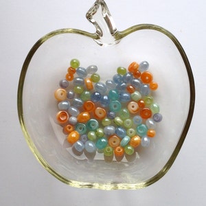 May include: A glass bowl shaped like an apple filled with colorful beads. The beads are in shades of blue, green, orange, and yellow.