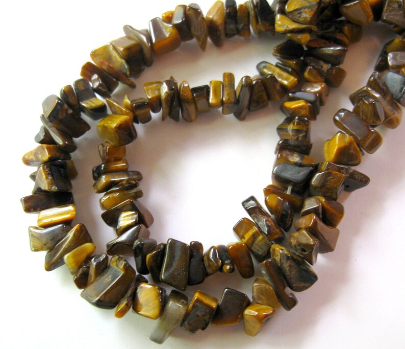 K&ouml;nnte beinhalten: Eine Str&auml;hne aus braunen und goldenen Tigerauge-Edelsteinkugeln. Die Kugeln sind unregelm&auml;&szlig;ig geformt und haben eine nat&uuml;rliche, raue Textur.
