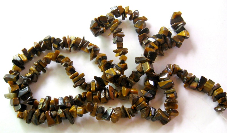 K&ouml;nnte beinhalten: Eine Str&auml;hne aus braunen und goldenen Tigerauge-Edelsteinkugeln. Die Kugeln sind unregelm&auml;&szlig;ig geformt und haben eine nat&uuml;rliche, raue Textur.