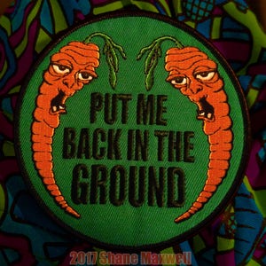 May include: A green embroidered patch with two orange carrots with faces. The carrots have a green stem and leaves. The text on the patch reads "Put me back in the ground" with the year "2017 Shane Maxwell" at the bottom.