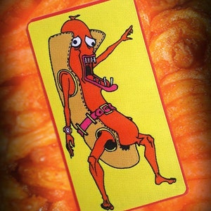 May include: A yellow and red embroidered patch featuring a cartoon hot dog with a human-like body and a surprised expression. The hot dog is wearing a brown bun and a pink belt with a buckle. The patch has a red border.