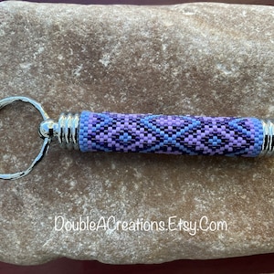 May include: A purple, blue, and pink beaded keychain with a silver key ring. The keychain is made of small beads and has a geometric pattern. The keychain is about 3 inches long.