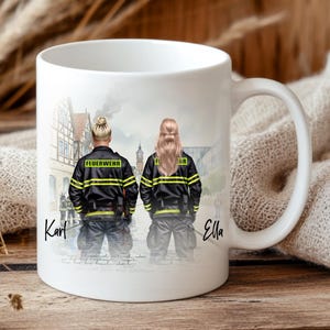 May include: White ceramic mug featuring a couple in firefighter uniforms, with the word "Feuerwehr" on their jackets. The mug has a handle and a detailed illustration of a town in the background. The names "Karl" and "Ella" are written on the mug.