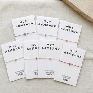 Könnte beinhalten: Eine Auswahl an bunten Armbändern mit Smiley-Perlen. Jedes Armband ist an einer weißen Karte mit der Aufschrift "MUT ARMBAND" und einem personalisierten Namen befestigt. Die Armbänder sind in verschiedenen Farben erhältlich.