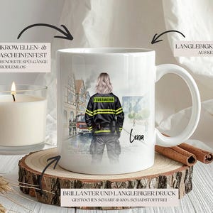 May include: White ceramic mug featuring a firefighter illustration. The mug has a handle and a printed design of a firefighter in uniform. The mug is on a wooden coaster with a lit candle and cinnamon sticks.