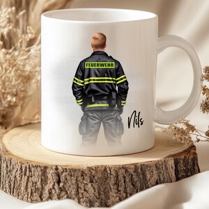 May include: White ceramic mug featuring a firefighter illustration. The firefighter is wearing a black jacket with yellow stripes and the word "FEUERWEHR". The mug is on a wooden slice, with the name "Nils" written on the mug.