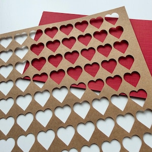 May include: A rectangular sheet of brown kraft paper featuring a repeating grid pattern of die-cut heart shapes. The sheet is layered over a solid red textured cardstock, creating a contrast between the brown paper and the red background.