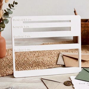 May include: A white mail size guide with four horizontal slots labeled with measurements in centimeters. The text includes "Standard 0.5 cm", "Kompakt 1 cm", "Großbriel 2 cm", and "Maxibrief 5 cm". The guide is on a textured surface with envelopes.