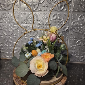 Könnte beinhalten: Oster-Blumenarrangement in einem goldenen Hasenohrenrahmen. Das Arrangement umfasst pastellfarbene Blumen, Kunst-Eier und Grünzeug, auf einer Holzbasis. Die Hasenohren sind aus Goldmetall.