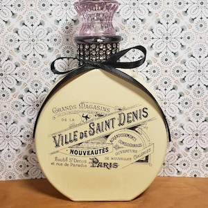 Könnte beinhalten: Eine dekorative, runde Flasche mit cremefarbenem Körper und schwarzem Band. Die Flasche hat einen rosa Glasverschluss und einen schwarzen Kragen mit Strasssteinen. Der Text auf der Flasche lautet "GRANDS MAGASINS DE LA VILLE DE SAINT DENIS PARIS."