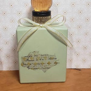 Pode incluir: Uma garrafa retangular verde claro com uma rolha esférica decorativa de cor âmbar. A garrafa é adornada com uma fita dourada e branca e um rótulo com texto manuscrito. A rolha assenta numa base preta com strass dourados.