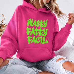 May include: A bright pink hooded sweatshirt featuring a front kangaroo pocket and lime green text printed on the chest that reads NASTY FATTY FACIL with a small outlined heart shape at the end of the final word in a bold graffiti style font.