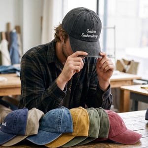 May include: A collection of baseball caps in various colors, including navy, tan, denim, mustard, olive, and burgundy. One cap is held up, displaying the text "Custom Embroidery". Another cap has the name "nah" embroidered on it.