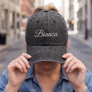May include: A black, distressed baseball cap with the name "Bianca" embroidered in white script. The cap is worn by a person in a denim jacket, with a blurred city street in the background.