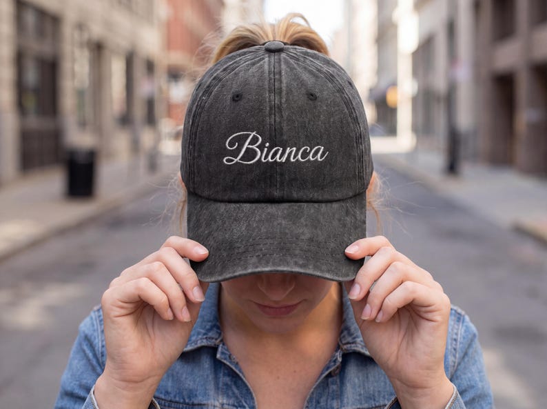 May include: A black, distressed baseball cap with the name "Bianca" embroidered in white script. The cap is worn by a person in a denim jacket, with a blurred city street in the background.