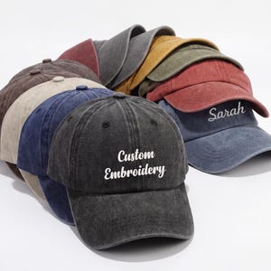 May include: A collection of baseball caps in various colors, including black, brown, blue, and red. One cap features the text "Custom Embroidery," while another has the name "Sarah." The caps are arranged in a stack, showcasing their different hues.