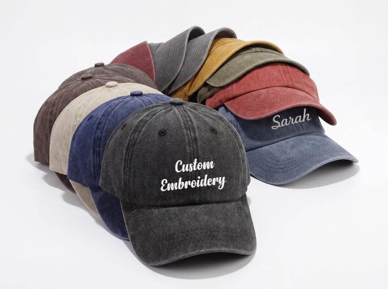 May include: A collection of baseball caps in various colors, including black, brown, blue, and red. One cap features the text "Custom Embroidery," while another has the name "Sarah." The caps are arranged in a stack, showcasing their different hues.