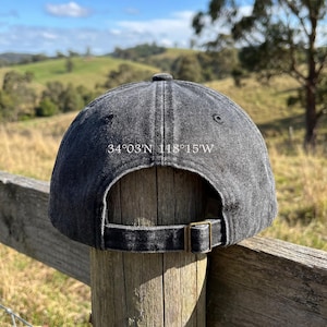 May include: A dark gray baseball cap with the coordinates 34&deg;03'N 118&deg;15'W printed on the back. The cap has a curved brim and an adjustable strap with a brass-colored buckle. The background shows a rural landscape.