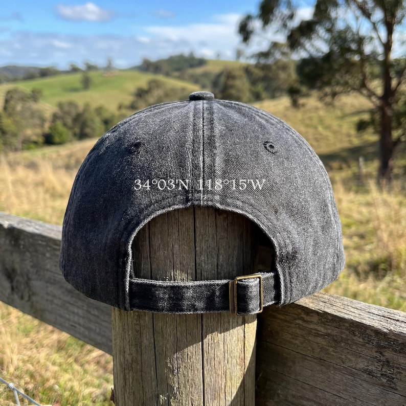 May include: A dark gray baseball cap with the coordinates 34&deg;03'N 118&deg;15'W printed on the back. The cap has a curved brim and an adjustable strap with a brass-colored buckle. The background shows a rural landscape.