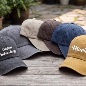 May include: A collection of six baseball caps in various colors, including gray, tan, brown, and blue, arranged on a wooden surface. One cap features the text "Custom Embroidery," and another displays the name "Maria."