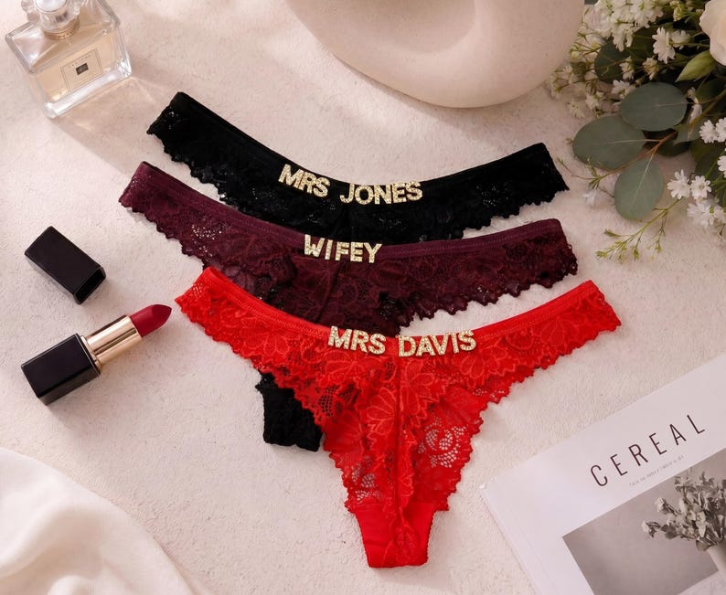 May include: Three pairs of lace thong underwear in black, burgundy, and red. Each pair features gold-toned rhinestone lettering on the front waistband. The black pair reads MRS JONES, the burgundy pair reads WIFEY, and the red pair reads MRS DAVIS.