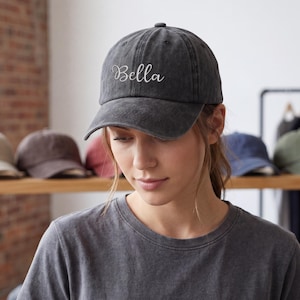 May include: A dark gray baseball cap with the name "Bella" embroidered in white script. The cap is worn by a person. Other caps in various colors are visible on a shelf in the background.