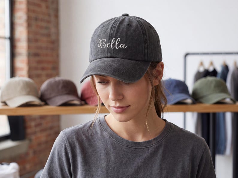 May include: A dark gray baseball cap with the name "Bella" embroidered in white script. The cap is worn by a person. Other caps in various colors are visible on a shelf in the background.