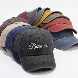 May include: A stack of baseball caps in various colors, including gray, brown, blue, and red. The front cap is dark gray with the name "Bianca" embroidered in white script. The caps have a worn, vintage look.